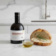 A bottle of Greek organic EVOO on a white marble kitchen counter next to sliced sourdough bread, a rosemary sprig, and a small bowl of golden unfiltered olive oil.