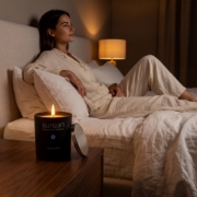A cozy bedroom atmosphere with the Aurean Nox candle lit on a wooden bedside table while a woman relaxes in linen pajamas, emphasizing a calming wellness routine.